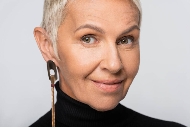 positive senior woman with long earring and black turtleneck isolated on grey - Fotoğraf, Görsel