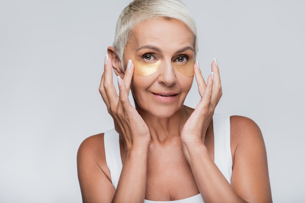 pleased senior woman applying collagen eye patches isolated on grey - Zdjęcie, obraz