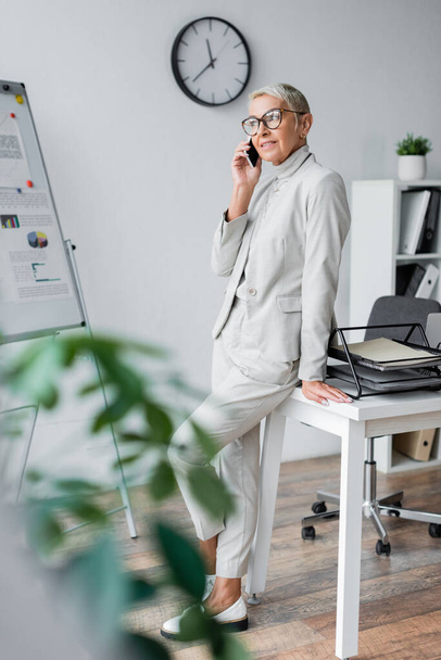 radosna starsza bizneswoman w okularach rozmawia przez telefon komórkowy w biurze  - Zdjęcie, obraz