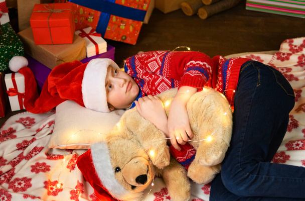 Παιδί κορίτσι κοιμάται στο νέο έτος ή χριστουγεννιάτικη διακόσμηση. Φώτα διακοπών και δώρα, χριστουγεννιάτικο δέντρο διακοσμημένο με παιχνίδια. Φοράει ένα κόκκινο πουλόβερ και ένα καπέλο βοηθού του Άη Βασίλη. - Φωτογραφία, εικόνα