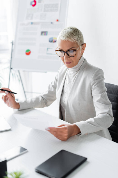 senior businesswoman in glasses looking at document in office - Фото, изображение