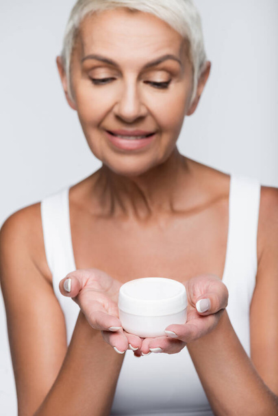 blurred and happy senior woman looking at container with cosmetic cream isolated on grey - Foto, Bild