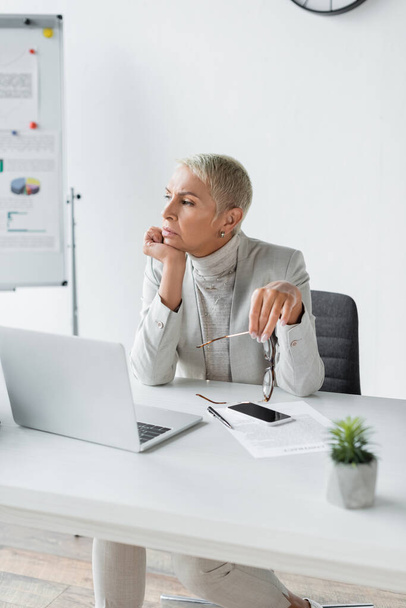 pensive businesswoman with grey hair holding glasses at workplace  - Valokuva, kuva