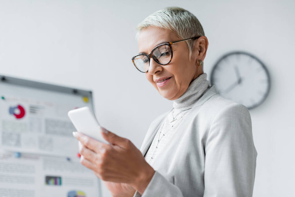pleased senior businesswoman in glasses looking at smartphone  - Photo, Image