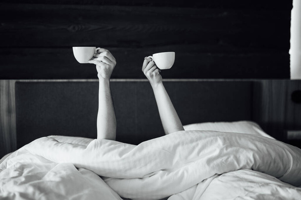 A pair of hands are holding over the blanket in the bed are holding cups of coffee. Morning time - Фото, зображення