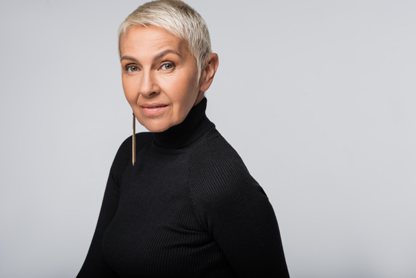 smiling senior woman with long earring and black turtleneck looking at camera isolated on grey - Фото, зображення