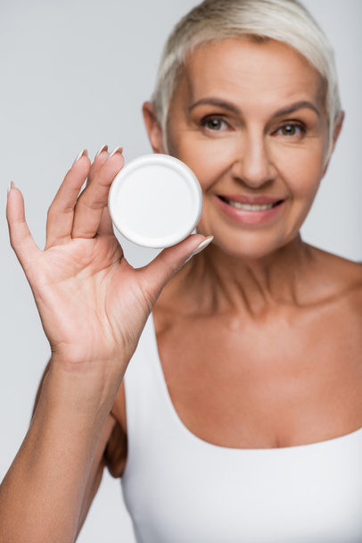 positive elderly woman holding container with cosmetic cream isolated on grey - Photo, Image