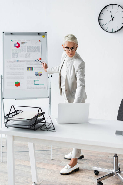 senior businesswoman in glasses looking at laptop while standing near flip chart  - Foto, Bild
