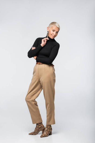 volle Länge der zufriedenen und stilvollen Seniorin in beiger Hose und Rollkragen posiert auf grau - Foto, Bild
