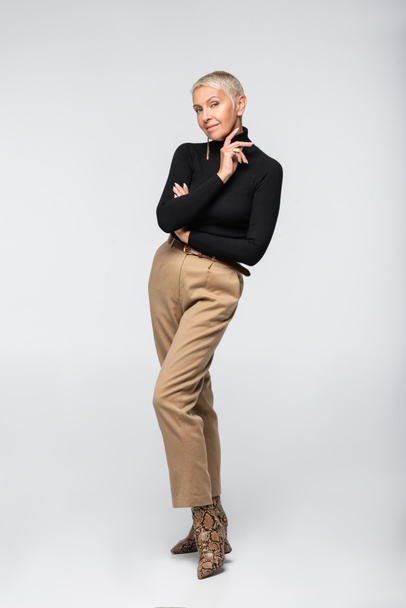 full length of pleased and trendy senior woman in beige pants and turtleneck posing on grey - Фото, зображення