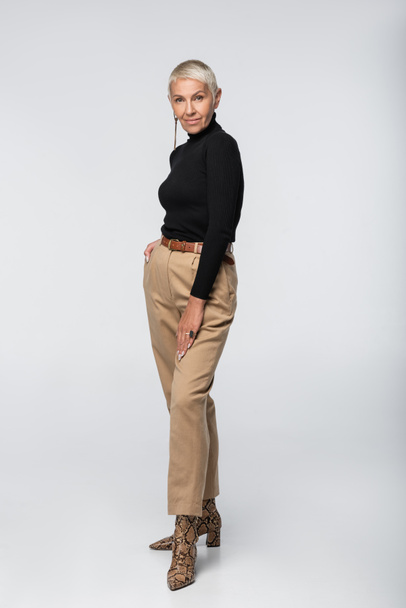 full length of trendy senior woman in black turtleneck and animal print boots posing on grey - Фото, зображення