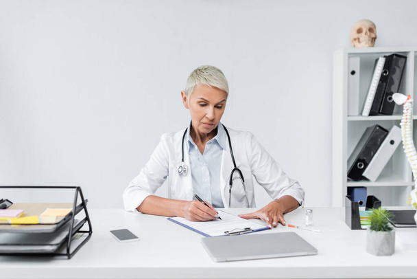 senior doctor with grey hair writing prescription near devices on desk  - Fotoğraf, Görsel