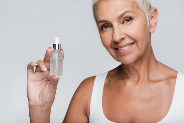happy elderly woman holding bottle with serum isolated on grey - Foto, imagen