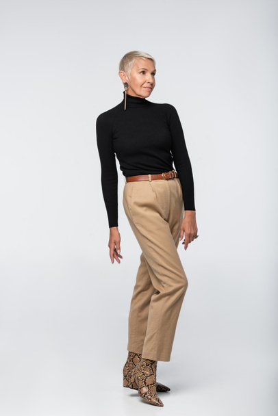 full length of stylish senior woman in beige pants, black turtleneck and animal print boots posing on grey - Foto, afbeelding
