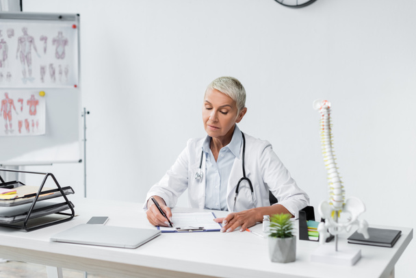 doctor with grey hair writing prescription near devices on desk  - 写真・画像
