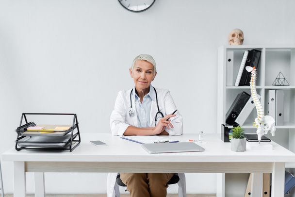Fröhlicher Oberarzt im weißen Kittel lächelt neben Geräten auf dem Schreibtisch  - Foto, Bild