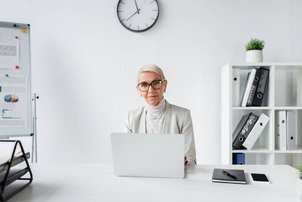 старшая деловая женщина в очках сидит за столом с устройствами в офисе  - Фото, изображение