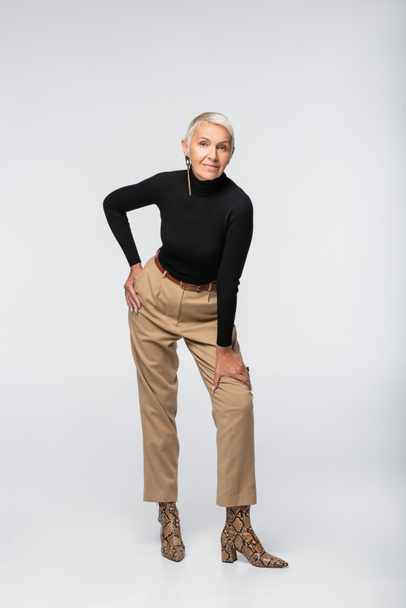 full length of pleased and trendy senior woman in beige pants, black turtleneck and animal print boots posing on grey - Foto, immagini
