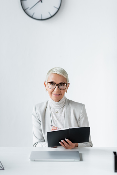 cheerful senior businesswoman in glasses writing in notebook near laptop - Foto, Imagem