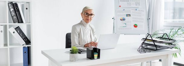 オフィスのデバイスやバナー付きのデスクに座っている眼鏡のシニアビジネス女性 - 写真・画像