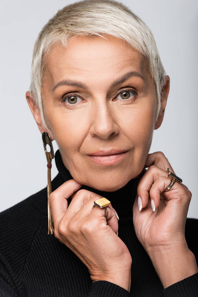 portrait of senior woman with long earring and black turtleneck isolated on grey - Photo, image