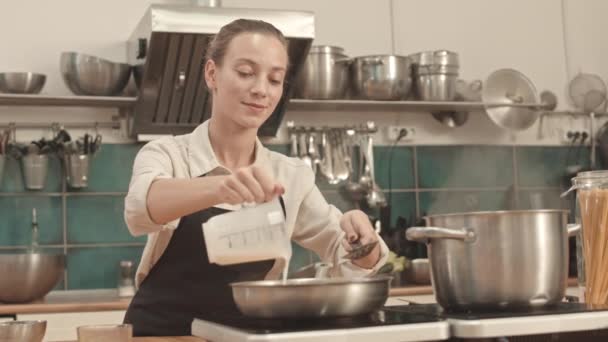 Средний низкий угол юной кавказской женщины в фартуке, стоящей у плиты на профессиональной кухне, добавляющей сливки в еду на сковородке и смешивающей - Кадры, видео