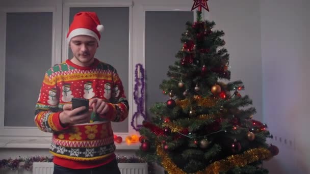 Een jongeman praat via video link en vertelt zijn familie over zichzelf, hij draagt een kersttrui en een hoed, naast hem staat een nieuwjaarsboom, een concept van Kerstmis en Nieuwjaar - Video