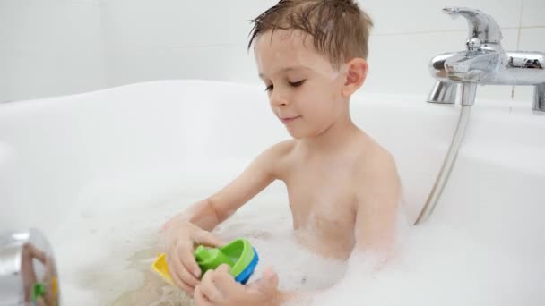 Cute boy sitting in bath with soap foam and playing toy boats and ships. Concept of family time, children development and fun at home - Footage, Video