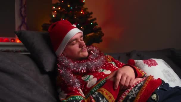 Młody człowiek śpi na kanapie, jest zmęczony, nosi świąteczny sweter i kapelusz, obok niego jest drzewo noworoczne, koncepcja Bożego Narodzenia i Nowego Roku - Materiał filmowy, wideo