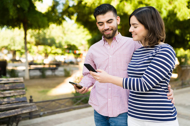 公園を歩いている間に彼女のボーイフレンドにスマートフォンでオンライン写真を示す幸せなガールフレンド - 写真・画像