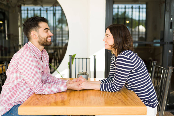 Side view of a beautiful couple in love gazing at each other and holding hands while sitting at a restaurant - Photo, Image