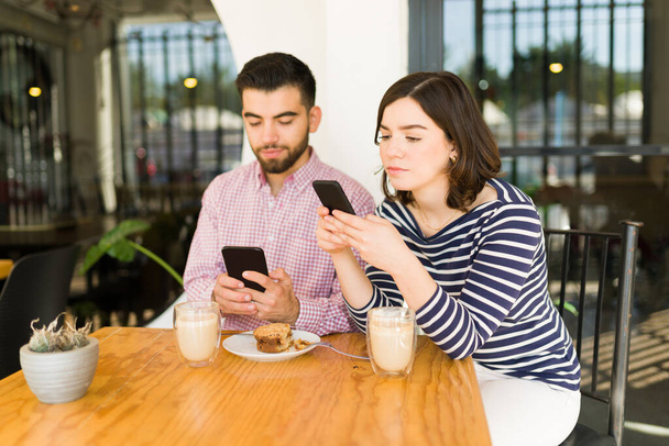 Znudzona para po dwudziestce, pisząca SMS-y i oglądająca media społecznościowe na telefonach podczas nudnej randki w kawiarni. - Zdjęcie, obraz
