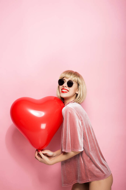 Sexy blonde hugging in the shape of a heart a red ball and smiling. On a pink background. - Φωτογραφία, εικόνα