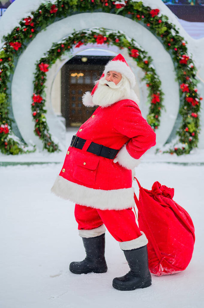 Santa Claus posing with a bag of gifts on the background of Christmas decorations outdoors - Photo, Image