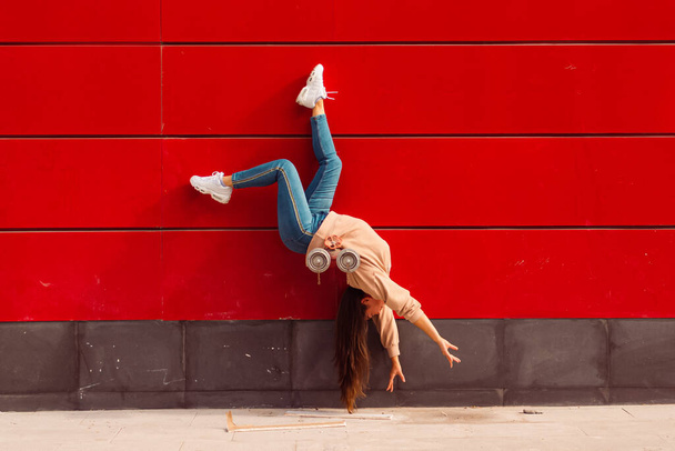 A gymnast in a twine on the street on a red background, in blue jeans and a beige sweater - Photo, Image