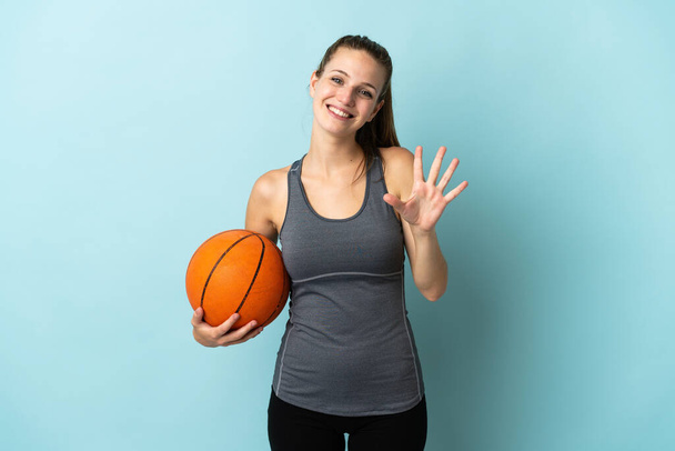Nuori nainen pelaa koripalloa eristetty sinisellä taustalla laskenta viisi sormilla - Valokuva, kuva