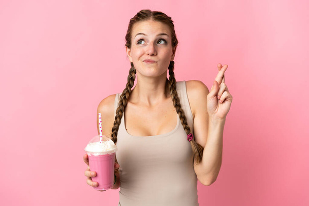 Молодая женщина с клубничным молочным коктейлем изолирована на розовом фоне с скрещиванием пальцев и желанием всего наилучшего - Фото, изображение