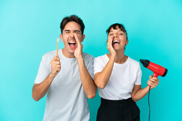 Joven pareja de raza mixta sosteniendo un secador de pelo y cepillo de dientes aislados sobre fondo azul gritando y anunciando algo - Foto, imagen