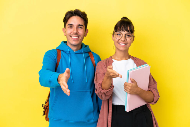 Young student couple isolated on yellow background shaking hands for closing a good deal - Foto, imagen