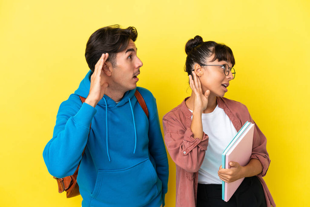 Young student couple isolated on yellow background listening to something by putting hand on the ear - Foto, Imagen