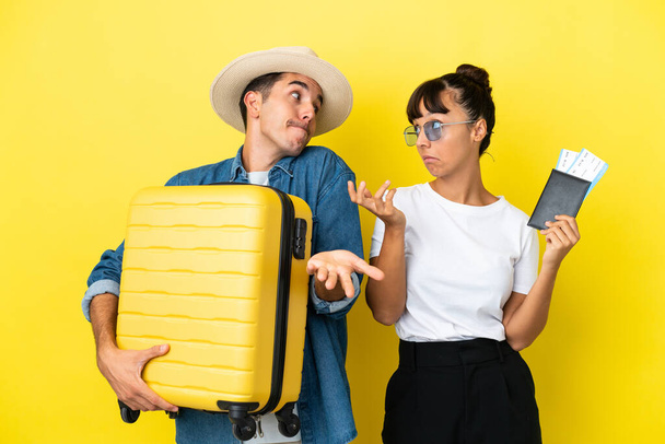 Молодые друзья-путешественники, держащие чемодан и паспорт изолированные на желтом фоне делают незначительный жест, поднимая плечи - Фото, изображение