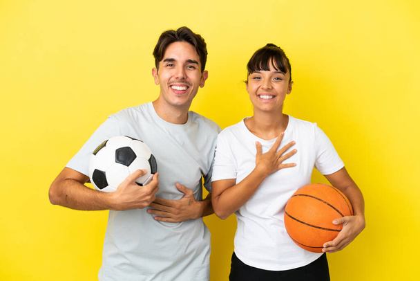 joven pareja deportiva jugando fútbol y baloncesto aislado sobre fondo amarillo sonriendo mucho mientras pone las manos en el pecho - Foto, imagen