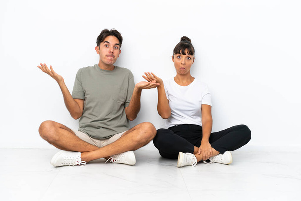 Young mixed race couple sitting on the floor isolated on white background having doubts while raising hands and shoulders - Photo, Image