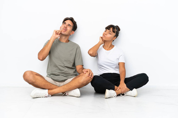 Young mixed race couple sitting on the floor isolated on white background thinking an idea while scratching head - Photo, Image