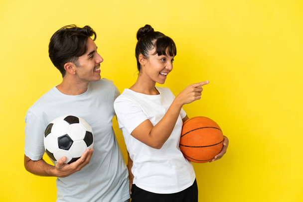 young sport couple playing football and basketball isolated on yellow background presenting an idea while looking smiling towards - Photo, Image