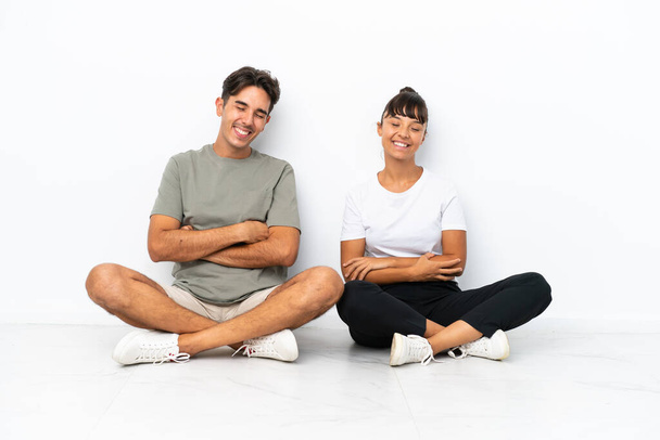 Young mixed race couple sitting on the floor isolated on white background keeping the arms crossed while smiling - Photo, Image