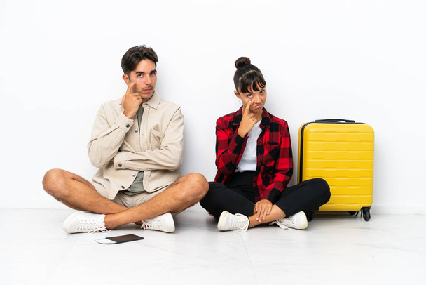 Giovani viaggiatori di razza mista coppia seduta sul pavimento isolato su sfondo bianco guardando davanti - Foto, immagini
