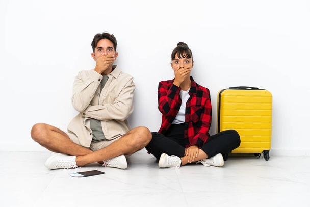 Молода змішана пара мандрівників, що сидять на підлозі ізольовані на білому тлі, покриваючи рот руками, щоб сказати щось недоречне
 - Фото, зображення