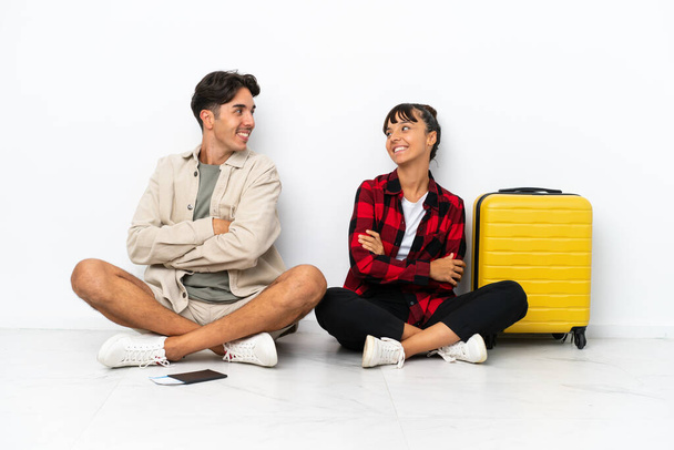 Giovani viaggiatori di razza mista coppia seduta sul pavimento isolata su sfondo bianco guardando sopra la spalla con un sorriso - Foto, immagini