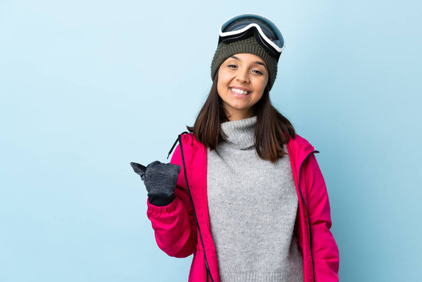 Menina esquiador de corrida mista com óculos de snowboard sobre fundo azul isolado apontando para o lado para apresentar um produto
. - Foto, Imagem